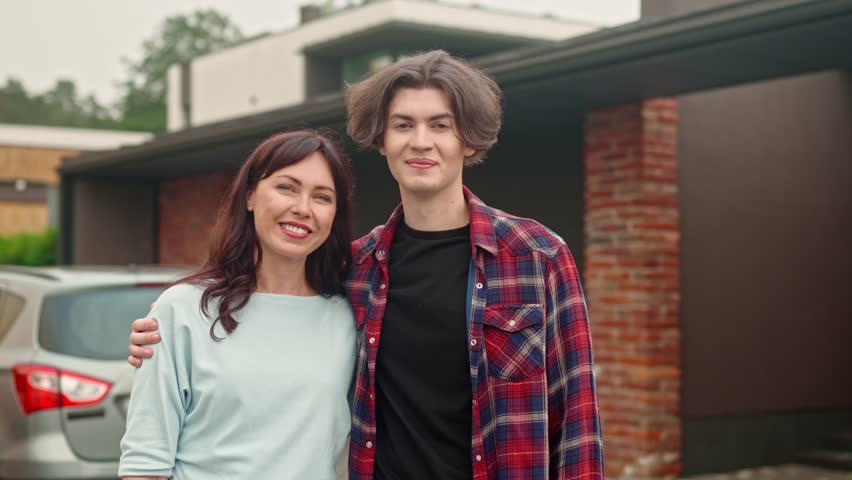 Pretty adult woman standing outside with young teenage guy. Mother and son are posing and smiling for camera on background of home yard. Good family relationship. Happy parenting concept.