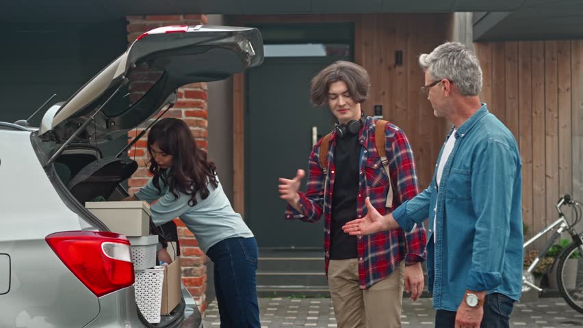 Parents help their son to put in moving boxes into car. Young boy entering university and says goodbye to his family. Dad shakes his son's hand. Leaving guy hugs mother. Happy family concept.