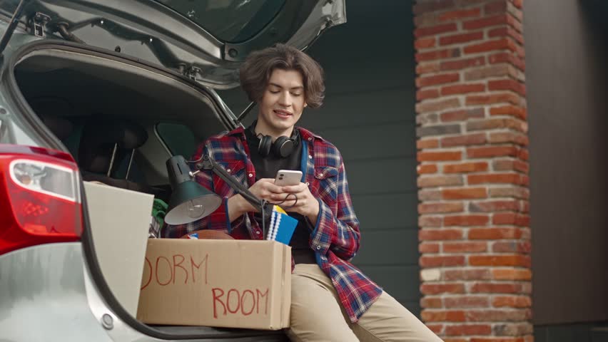 Close-up young male entering college and loading cardboard boxes into car. Adult boy sitting on car trunk and using phone for texting messages to his friends. Independent adult life. Relocation.