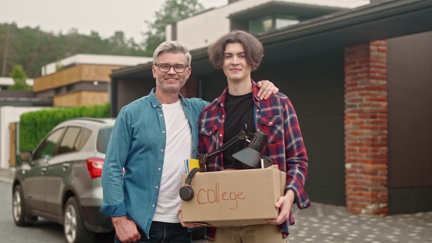 Portrait of adult man and young boy standing outside and looking at camera with smile. Cute guy entering college and moving away from home. Dad hugs his son leaving parents' house.