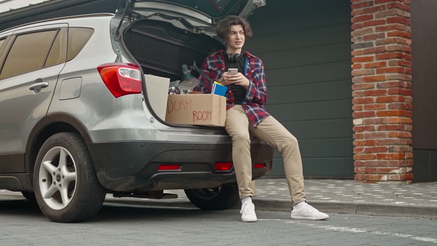 Young man entering college and loading cardboard boxes into car. Adult boy sitting on car trunk and using phone for texting messages to his friends. Teenager moving out of parent's home.