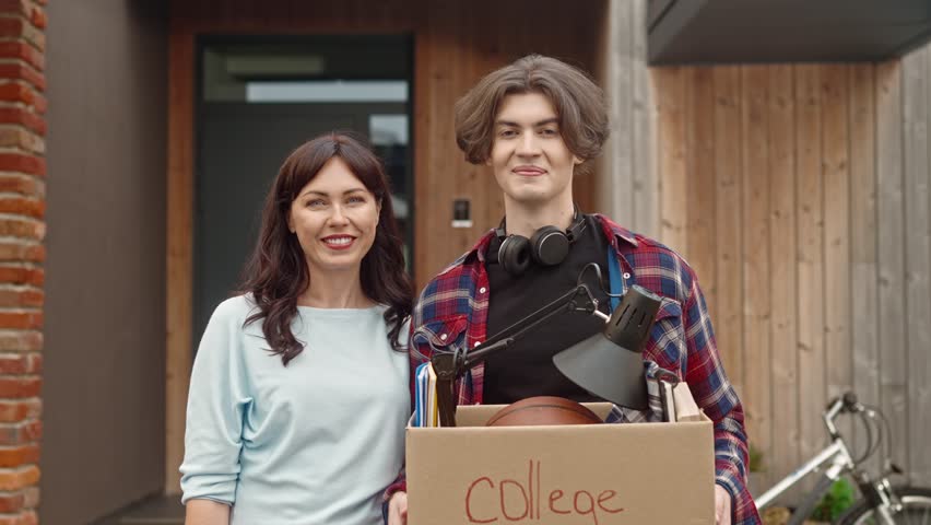Portrait of adult preaty woman and young boy standing outside and looking at camera with smile. Mother hugs her son leaving parents' house. Cute guy entering university and moving away from home.