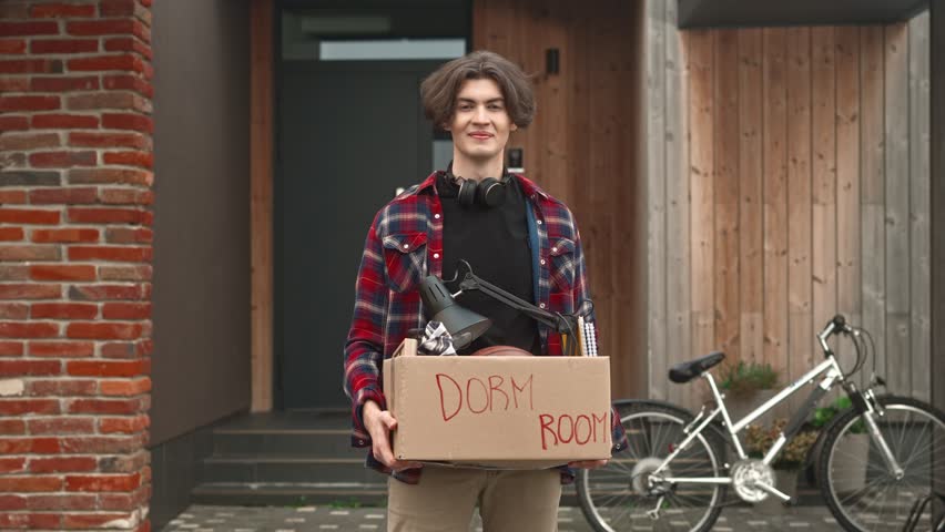 Portrait of handsome guy with headphones standing outside, looking at camera and posind. Young adult boy entering university and moving out of parent's home. Independent adult life. Relocation.