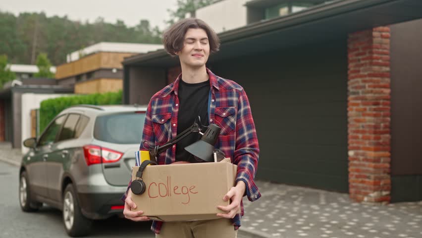 Portrait of handsome guy holding luggage box and standing outside. Handsome adolescent boy looking at camera smiling and posind. Tenager entering collegeand moving out of parent's home. Relocation.