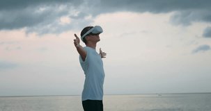 Young man sitting outdoors near the sea, wearing Virtual Reality headset, man spread his hands and enjoys a virtual world in virtual reality glasses, standing against the background of the sea
