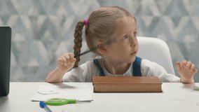 close up slow motion portrait of little girl student with pigtails in sundress, sitting at table at home, whiting, knocking pencil on book, nervous, kid looking for object with her eyes