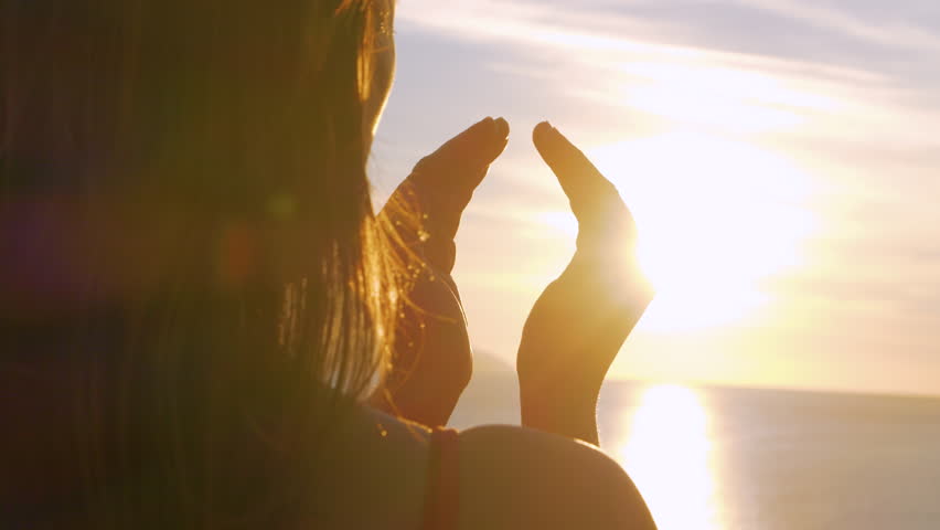 LENS FLARE, CLOSE UP: Golden sun rays shine between the palms of a young lady. She is trying to catch a glare of warm summer sun setting over blue Adriatic Sea while gentle breeze ruffles her hair. Royalty-Free Stock Footage #1104836847