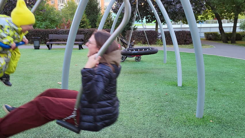 A happy child rides together with his mother on a swing. Toddler baby and his mom swing on the playground. Kid aged one year two months (14 months)