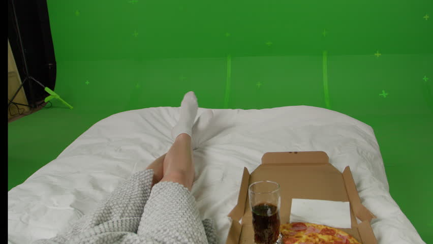 Back view of Woman isolated at home and eating pizza. The sick girl took a day off and relaxes lying on the bed and watching TV. Green screen chroma key background