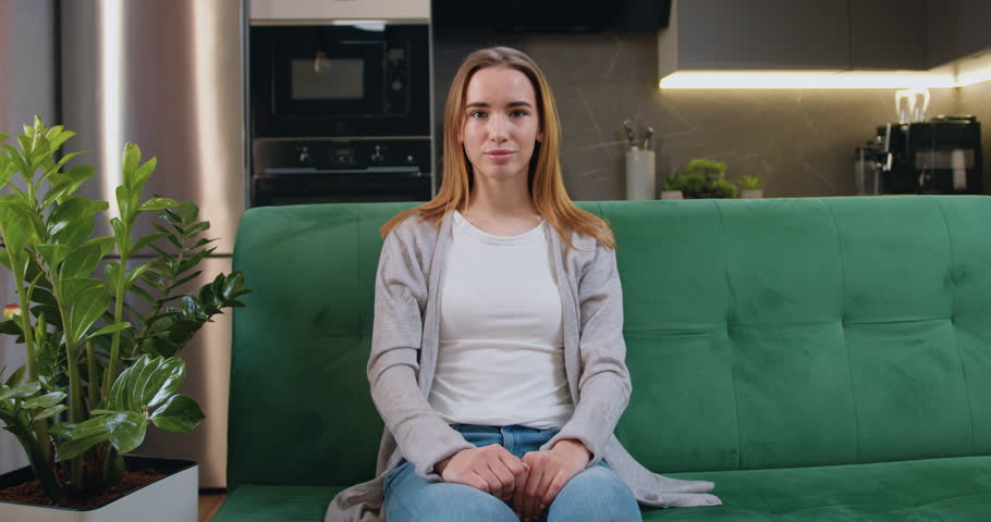 Smiling beautiful girl smiling while sitting on sofa and showing thumb up. Satisfied caucasian woman looking at camera with thumbs up sitting on a couch at home