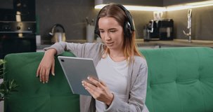 Beautiful young caucasian woman in casual clothes with headphones use a digital tablet and smiling while sitting on couch at kitchen background