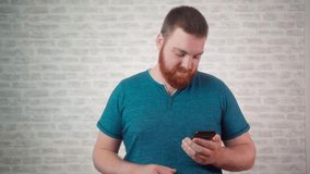 Handsome red bearded young guy 20s wearing green t-shirt isolated over grey background in studio. People sincere emotions lifestyle concept. Looking surprised wow, using mobile cell phone