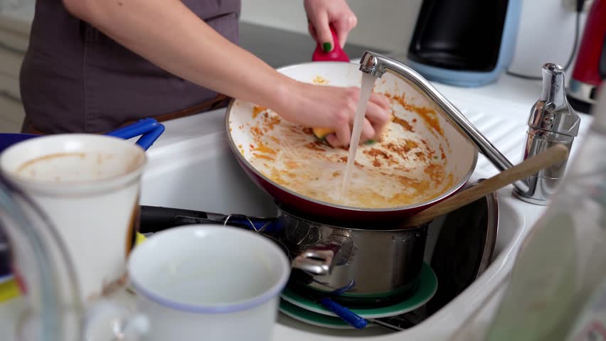 young woman in brown apron washes burnt frying pan with washcloth and dish detergent in kitchen sink. Nearby there is pile of dirty dishes on countertop. Royalty-Free Stock Footage #1104844943