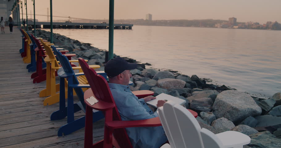 Man Using Laptop Outdoors While Sitting on a Wooden Chair by the Ocean in the City. Young Guy Works Remotely at a Computer on the Street. Remote Work, Business. | Shutterstock HD Video #1104846173