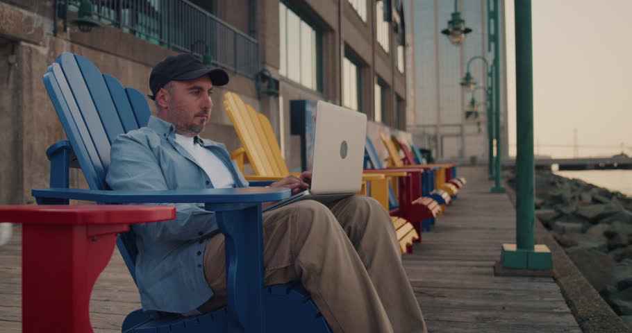 Man Using Laptop Outdoors While Sitting on a Wooden Chair by the Ocean in the City. Young Guy Works Remotely at a Computer on the Street. Remote Work, Business. | Shutterstock HD Video #1104846175