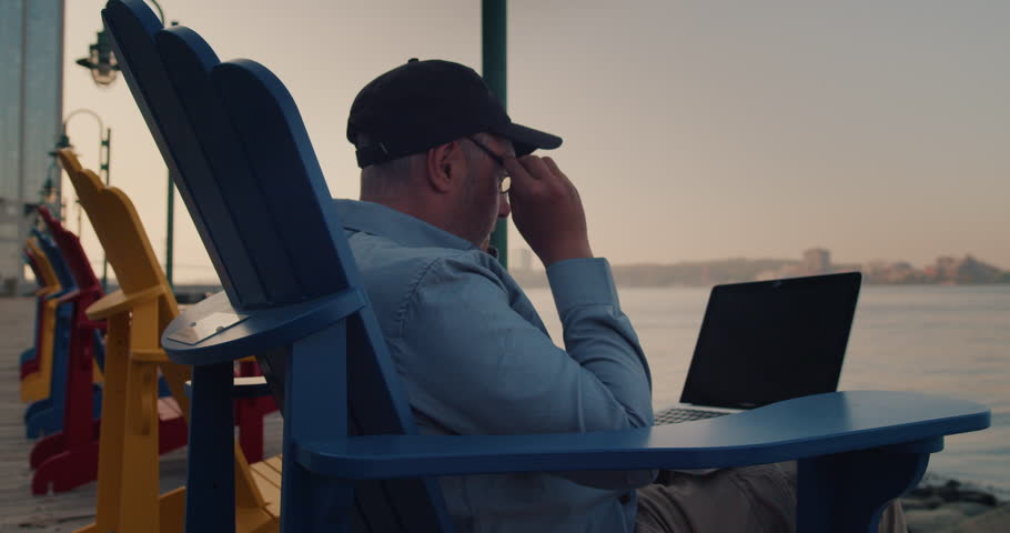 Man Using Laptop Outdoors While Sitting on a Wooden Chair by the Ocean in the City. Young Guy Works Remotely at a Computer on the Street. Remote Work, Business. | Shutterstock HD Video #1104846177
