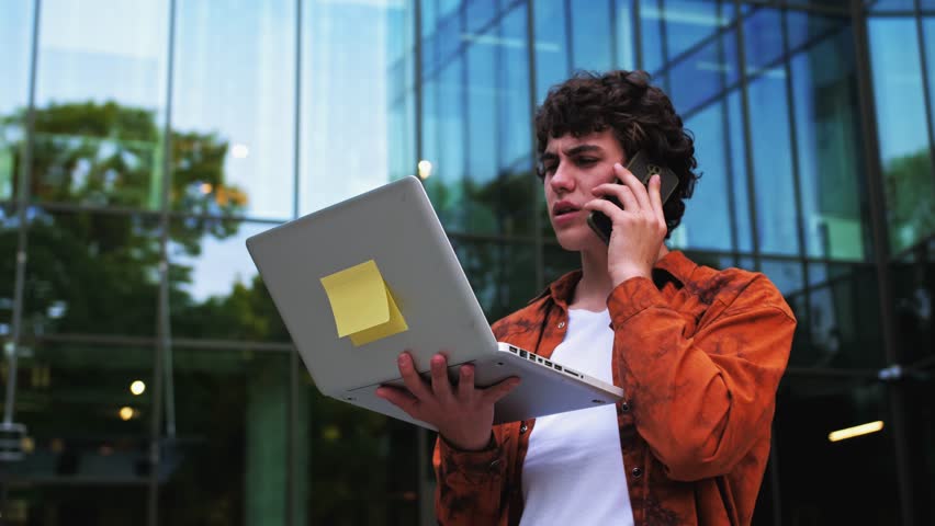  Man talking on cell phone pacing back and forth outdoors near office with laptop in his hands. Businessman working on pc and thinking. Online and remote work concept