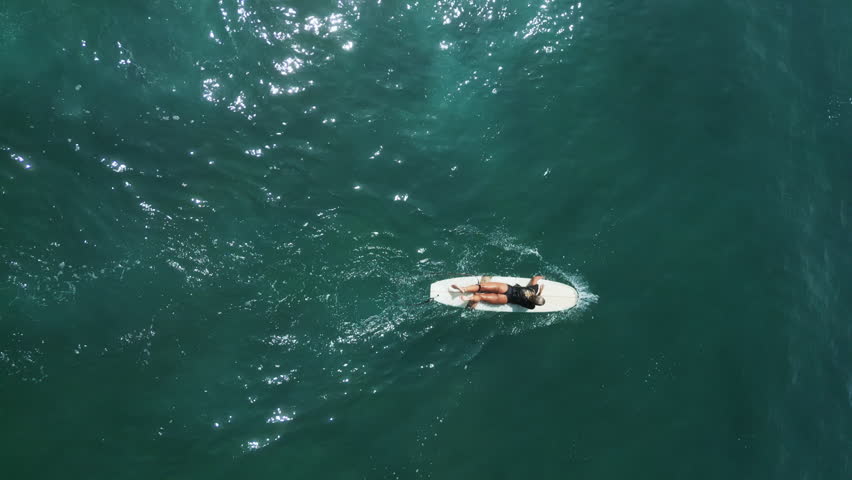 Young female surfer rowing on surfboard at scenic tropical location in sea on sunny day aerial shot. Surfing girl enjoying water sport paddling and surfing on summer vacation view from drone | Shutterstock HD Video #1104847241