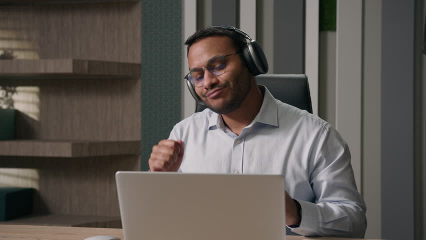 Excited happy funny carefree American businessman Indian man worker in wireless headphones having fun listen music with laptop computer audio sound app dance at office table dancing rhythm work break Royalty-Free Stock Footage #1104849933