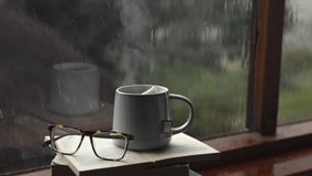 cinematic aesthetic cinematic relaxing therapy video after reading a book with a cup of hot coffee or tea steaming smoky with a book and glasses in the rain seen from the window
