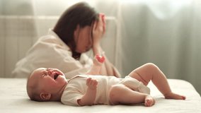 Tired infant girl cries on double bed against depressed mother sitting on floor. Baby daughter wants to eat getting parent attention - Powered by Shutterstock - Get 15% off with code: PIKWIZARD15