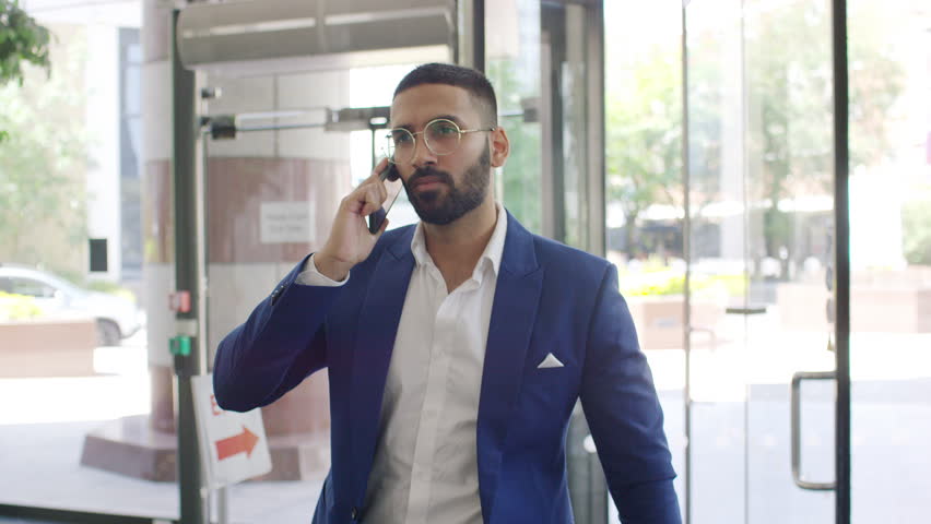 Handsome Indian male professional walks through a building lobby whilst talking on his phone, in slow motion Royalty-Free Stock Footage #1104863837