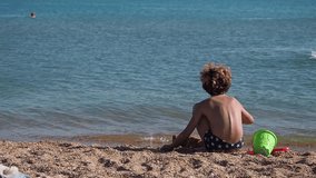 Small boy in blue swimwear plays with red shovel while sitting on sandy shore. Back view. Empty space for text