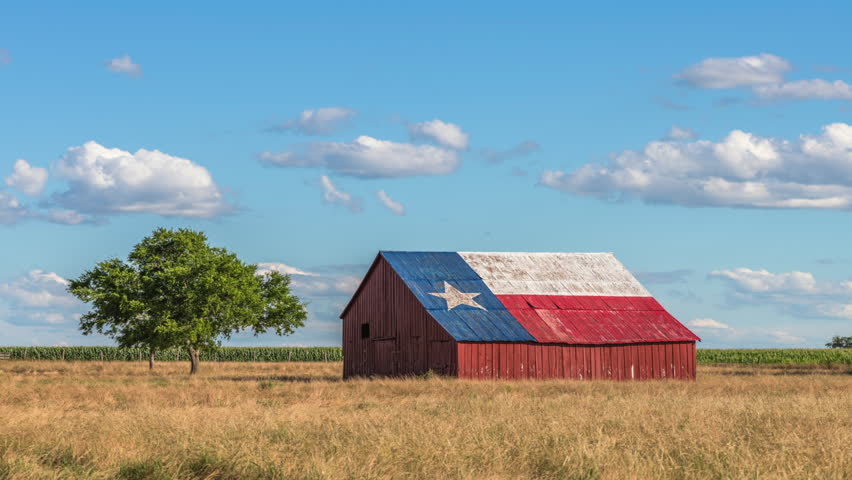 Time lapse footage of an abandoned old barn with the symbol of Texas painted on the roof sits in a rural area of the state, framed by farmland.