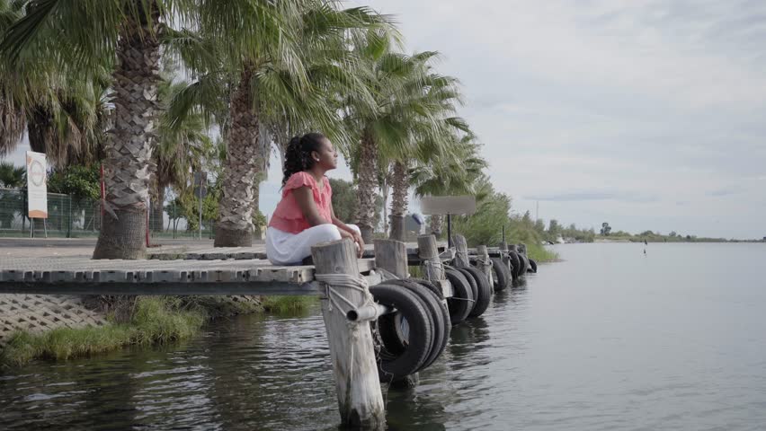Woman meditating by the river in a tropical landscape. Girl enjoying nature in a beautiful place sitting. Calm scene of an African American person disconnecting close to a lake. Digital detox concept. Royalty-Free Stock Footage #1104872799