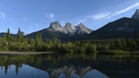 Timelapse sunset video of Three Sisters view seen in Canmore outside of Banff National Park with stunning reflection in water below. Clouds moving across sky with colours, blue sky, mountain peaks.