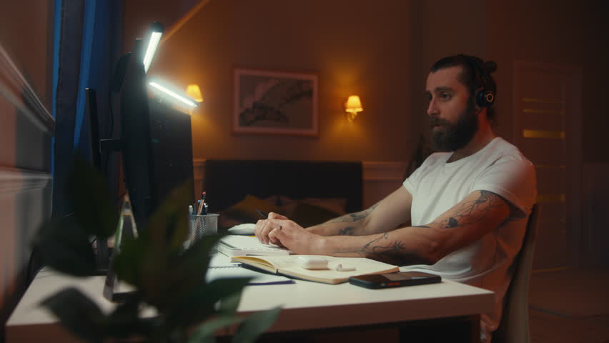 Concentrated financial analyst or trader with pen in hand types keyboard and makes notes. Man in headset works remotely in room late night. Table illuminated by LED lamps. Working at home concept.
