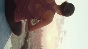 VERTICAL VIDEO: Young woman in bodysuit practices yoga at sunrise at viewpoint. Girl raises her hands up while sitting in lotus position. Back view