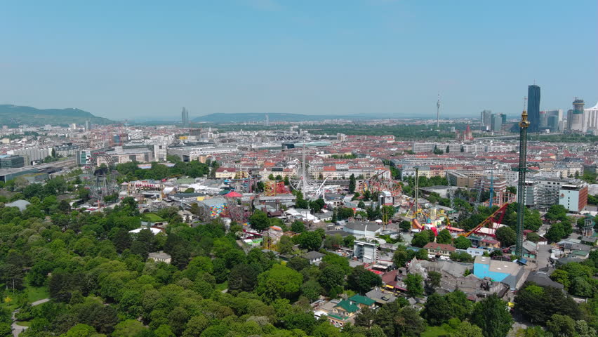 Aerial city view of Vienna, Austria, from above at Prater amusement park. Flying by iconic fairy wheel and other amusement rides ft beautiful Vienna skyline in background on a sunny summer day 5.5K