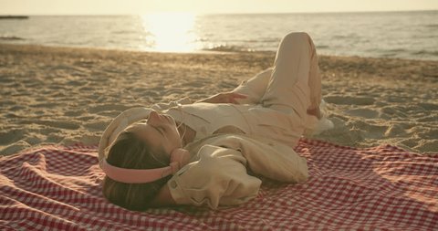 A blonde girl in light clothes and pink headphones listens to music and lies on a package on a sunny beach in the morning 스톡 비디오