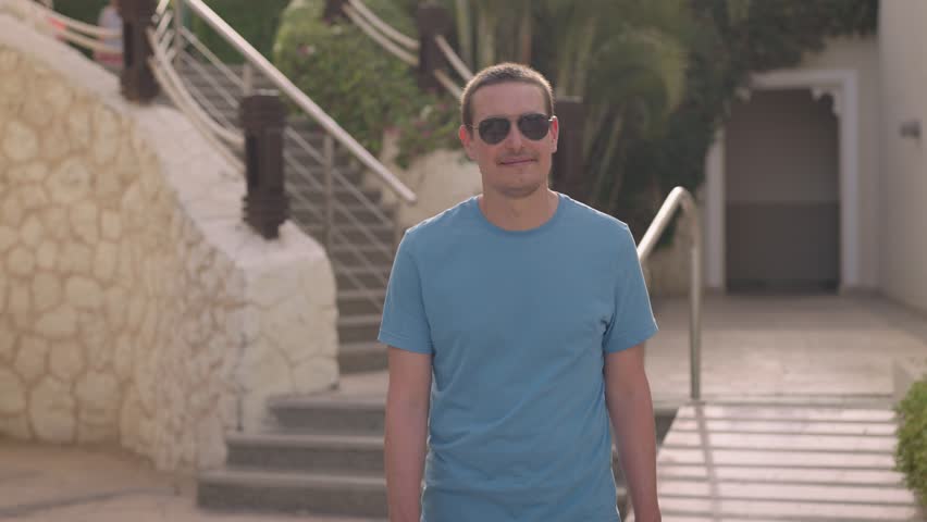 A young and successful man, wearing sunglasses, walks along the beautiful grounds of an expensive luxury resort. It