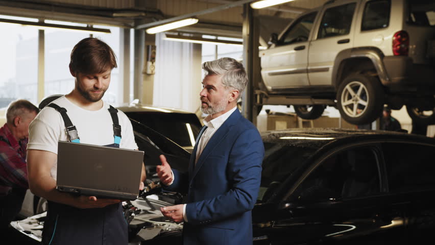 Handsome male mechanic wearing uniform, using laptop talking with grey hair business man client, finish repairing car or automobile components, working in car maintenance service center or shop