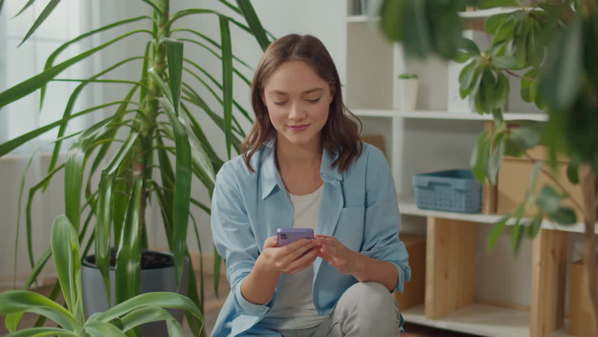 Smiling Young Woman Typing a Message on the Phone among Indoor Plants, Beautiful Young Woman with Beautiful Flowers, a Woman is Holding a Smartphone in her Hands, Stress-Free Life Thanks to Plants Royalty-Free Stock Footage #1104906569