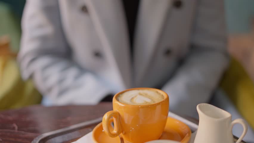 Closeup of woman holding, drinking coffee. Cafe, blue coat, orange coffee cup.