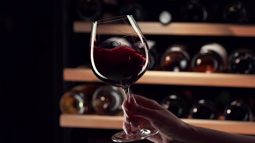 Close up female hand swirling red wine in wine glass. Wine expert tasting, rating and drinking wine, bottles in background. Slow motion video. Royalty-Free Stock Footage #1104910385