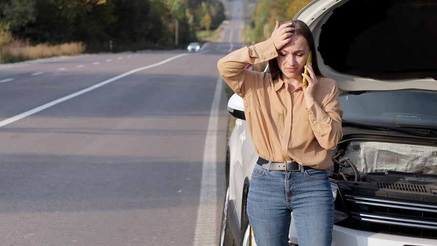 Upset young woman driver talking angrily on cell phone with assistance service near a broken car with open hood while inspecting engine having trouble with her vehicle Royalty-Free Stock Footage #1104910945