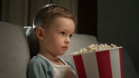 Toddler boy watches film for kids at movie projector and relaxes eating popcorn