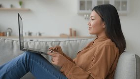 Asian relaxed woman girl on sofa couch at home kitchen talk laptop video meeting online conference talking with friend internet communication chinese businesswoman student smiling speaking side view