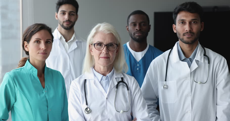 Mature female chief physician pose with young perspective multinational interns, wear uniform, smile look at camera feel satisfied with teamwork, career and professional advance. Medical council event Royalty-Free Stock Footage #1104911931