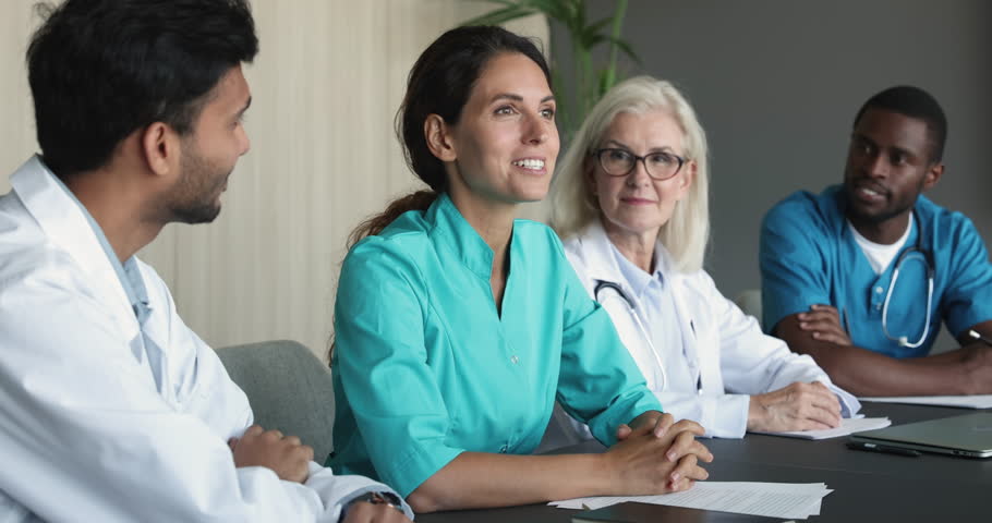 Attractive Hispanic woman physician and multinational colleagues, diverse workmates in coats participate group meeting event in clinic office boardroom. Medical council, share opinion about treatment Royalty-Free Stock Footage #1104911961