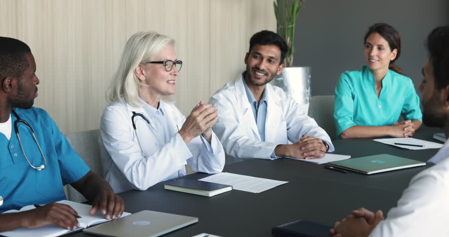 Elderly female chief physician makes speech during meeting with multiethnic colleagues or young, perspective interns. Diverse medicine employees in uniforms take part in medical council led by aged GP Royalty-Free Stock Footage #1104911971