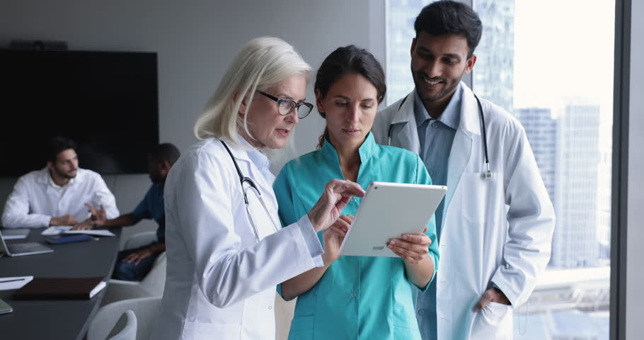 Mature and young clinic staff members, diverse professional GP therapists in uniforms discuss new medical application on digital tablet standing in conference room. Teamwork, modern tech, health-care Royalty-Free Stock Footage #1104911991
