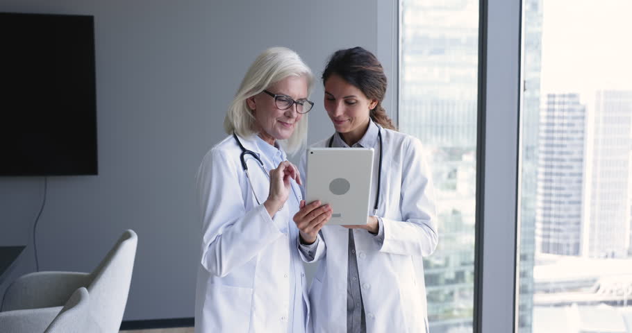 Young and mature female physicians talking standing in clinic office, discuss patient treatment plan, disease and symptoms using modern device tablet. Teamwork of intern and medical chief in hospital Royalty-Free Stock Footage #1104912011