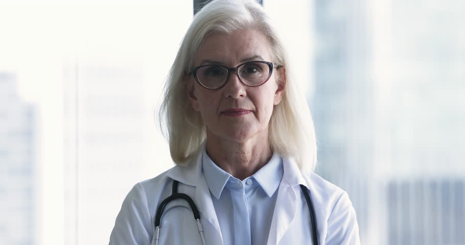 Close up head shot smiling mature 65s female general practitioner looking at camera, posing in clinic office. Portrait of cardiologist in white coat. Career and professional occupation, health-care Royalty-Free Stock Footage #1104912015