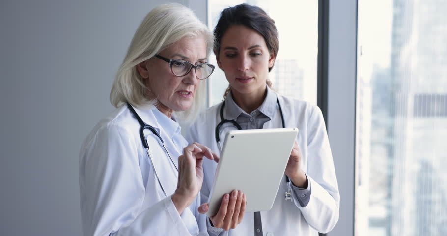 Women therapists, colleagues in white uniform with stethoscope on shoulder engaged in teamwork, discuss in hospital office use digital tablet app modern healthcare tech, review patient medical history Royalty-Free Stock Footage #1104912017