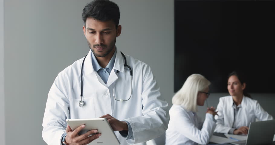 Focused young male Indian physician in white uniform holds digital tablet, use application for planning patient appointment in hospital office, check health history data, using on-line healthcare app Royalty-Free Stock Footage #1104912035